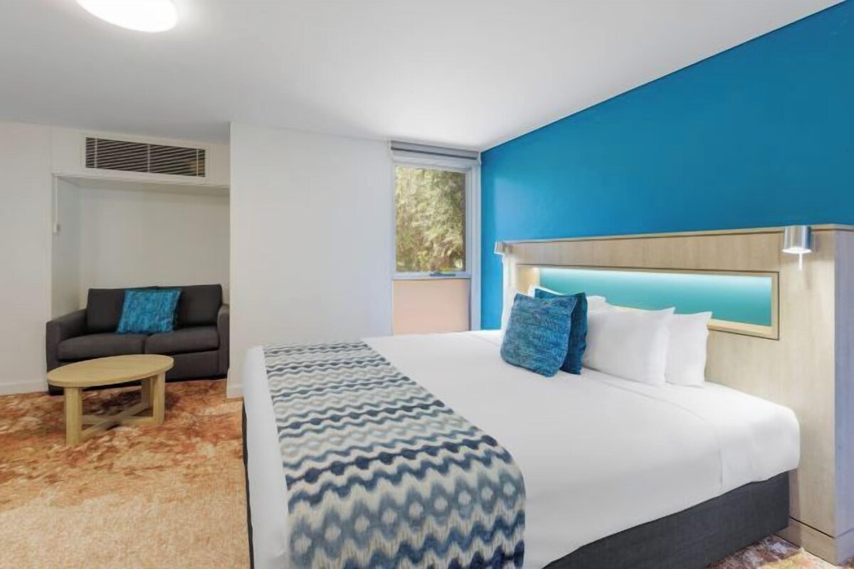Quarto de hotel com uma cama, um sofá, uma mesinha e uma janela.