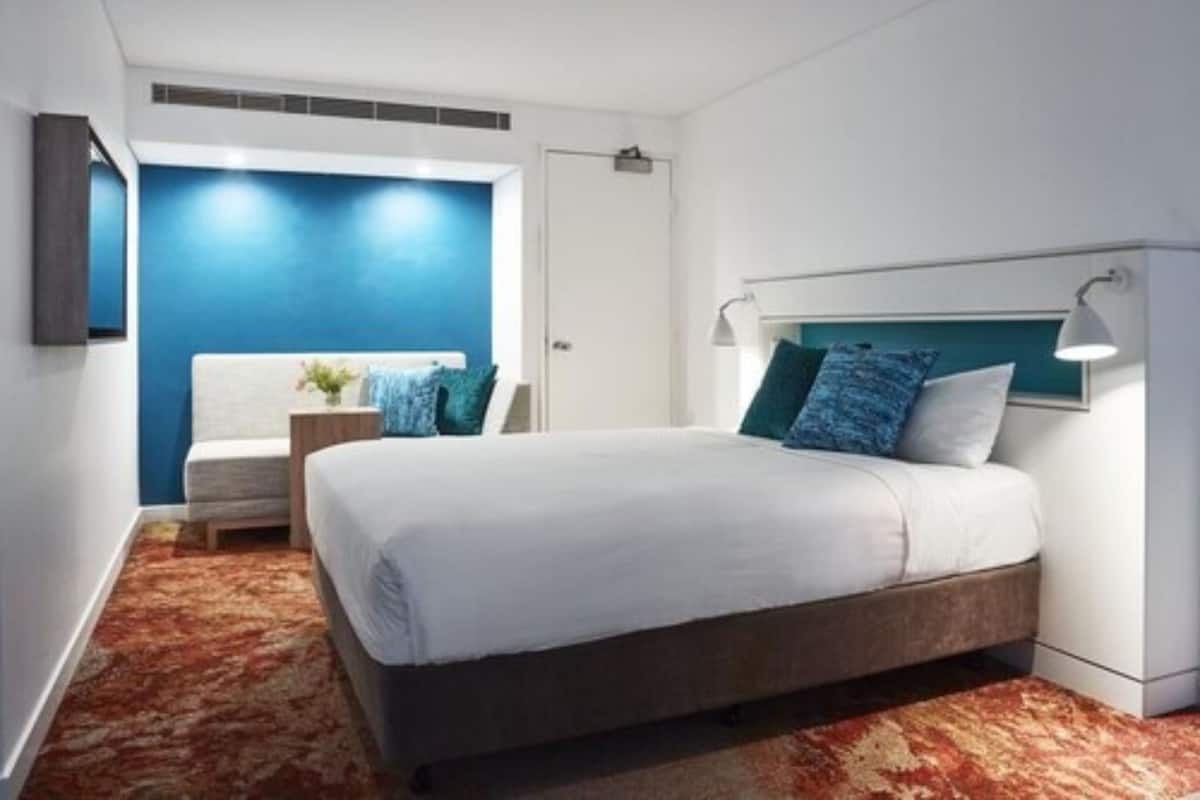 Una habitación de hotel moderna con una cama grande, una mesita de noche, una silla y una pared azul decorativa.