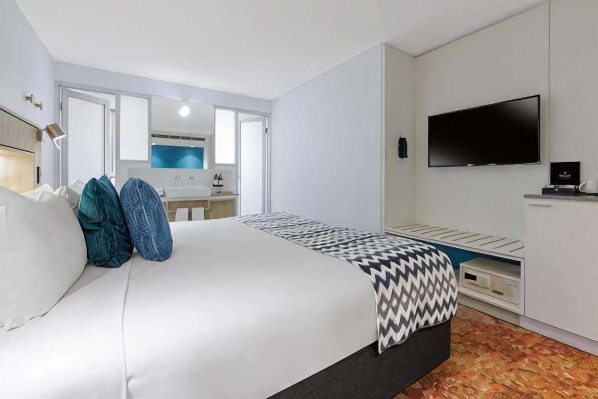 Habitación de hotel con cama, televisión, escritorio y baño visible a través de una puerta abierta.
