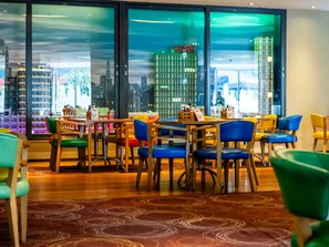 A dining area with tables and chairs, large windows with a cityscape view, and a patterned carpet. - Legoland Windsor Resort Hotel (Windsor)