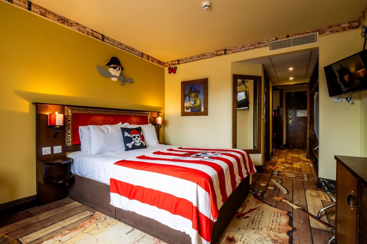 A hotel room with a large bed, a headboard with a skull and crossbones design, a red and white striped bedspread, a wooden floor, and a television mounted on the wall.