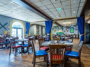 A spacious restaurant with wooden tables and chairs, a high ceiling with grid lights, and a stained glass window. - Legoland Windsor Resort Hotel (Windsor)