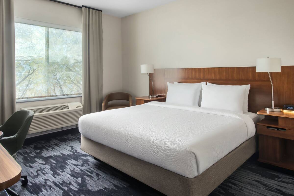 A hotel room with a large bed, two bedside tables, a chair, a desk, a window with curtains, and a view of trees.