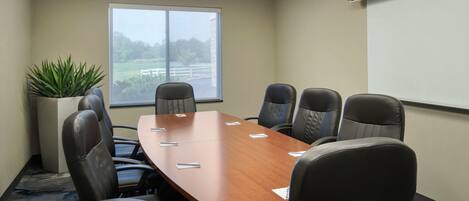A conference room with a long table, eight chairs, a projector screen, and a large window with a view of greenery.