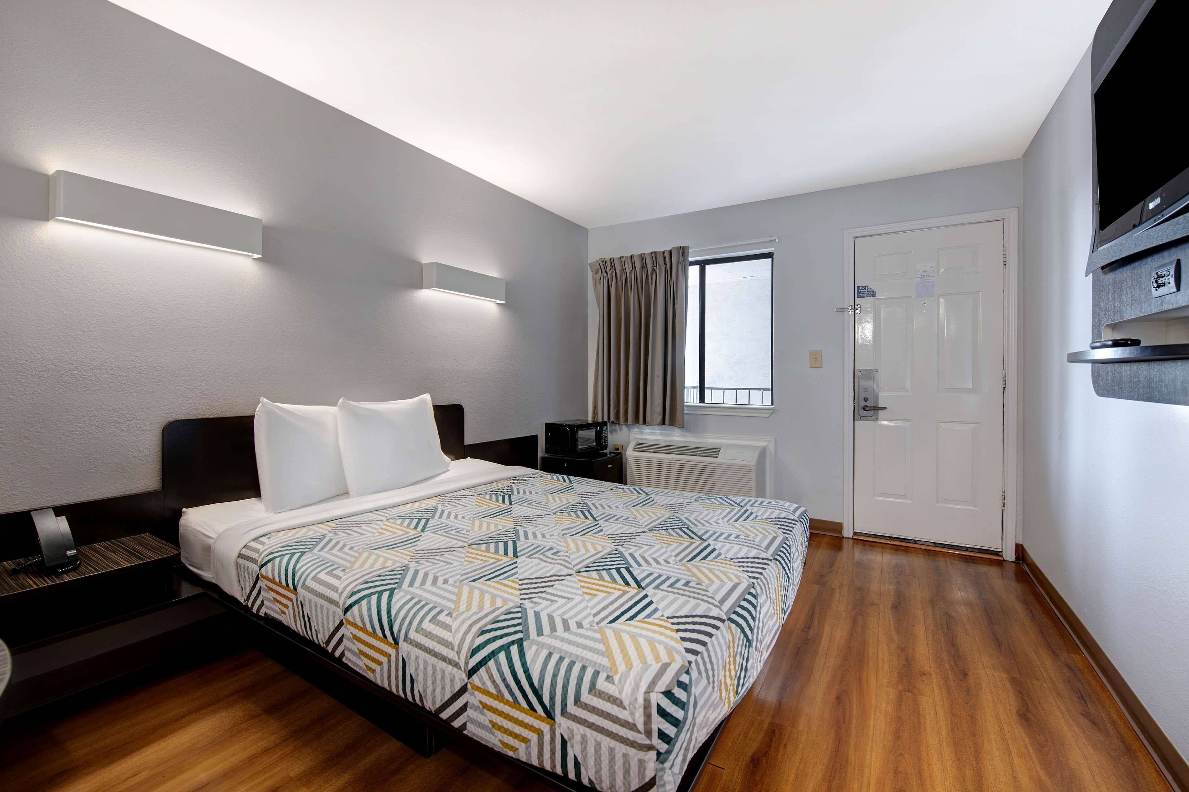 A hotel room with a bed, a desk, a flat-screen TV, a window with curtains, and a door.