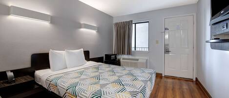 A hotel room with a bed, a desk, a flat-screen TV, a window with curtains, and a door.
