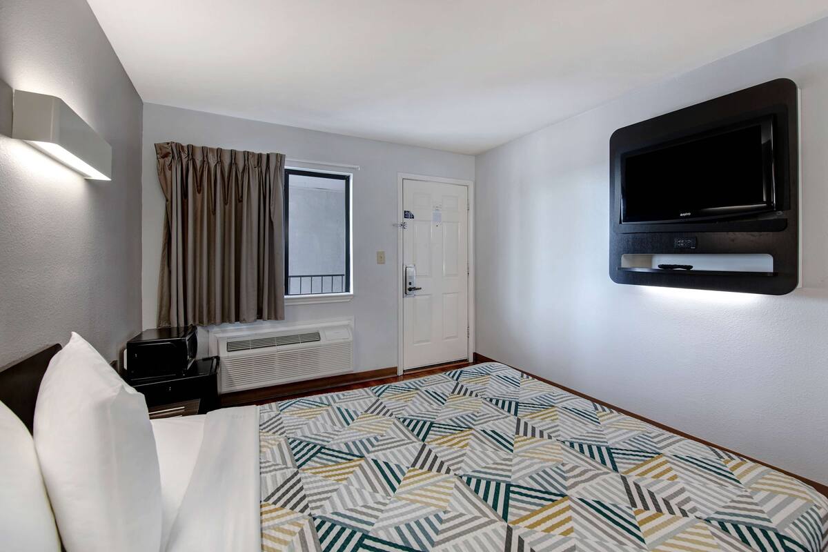 A hotel room with a bed, a flat-screen TV mounted on the wall, a window with curtains, a heater, and a small table with a lamp.