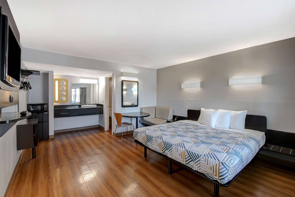 A modern hotel room with a bed, a desk, a chair, a small table, and a kitchenette.