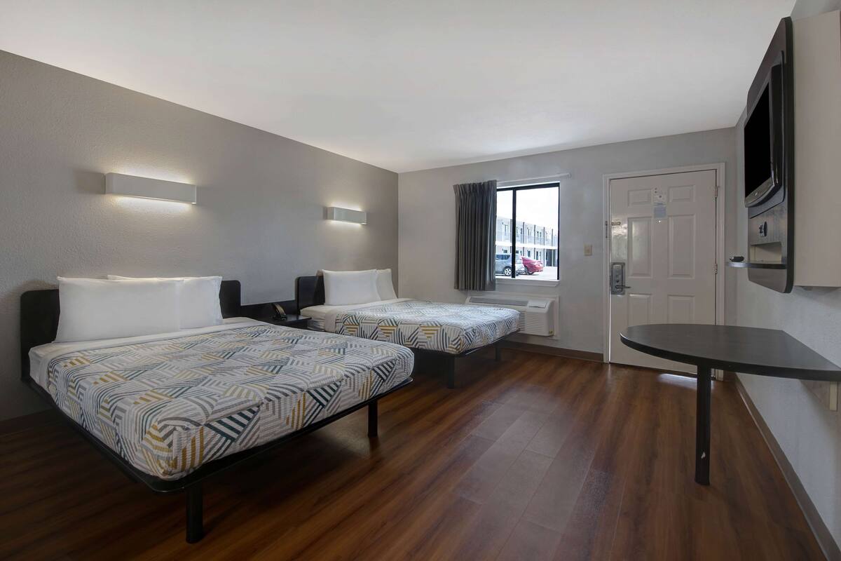 A hotel room with two beds, a small table, a TV, and a window with curtains.