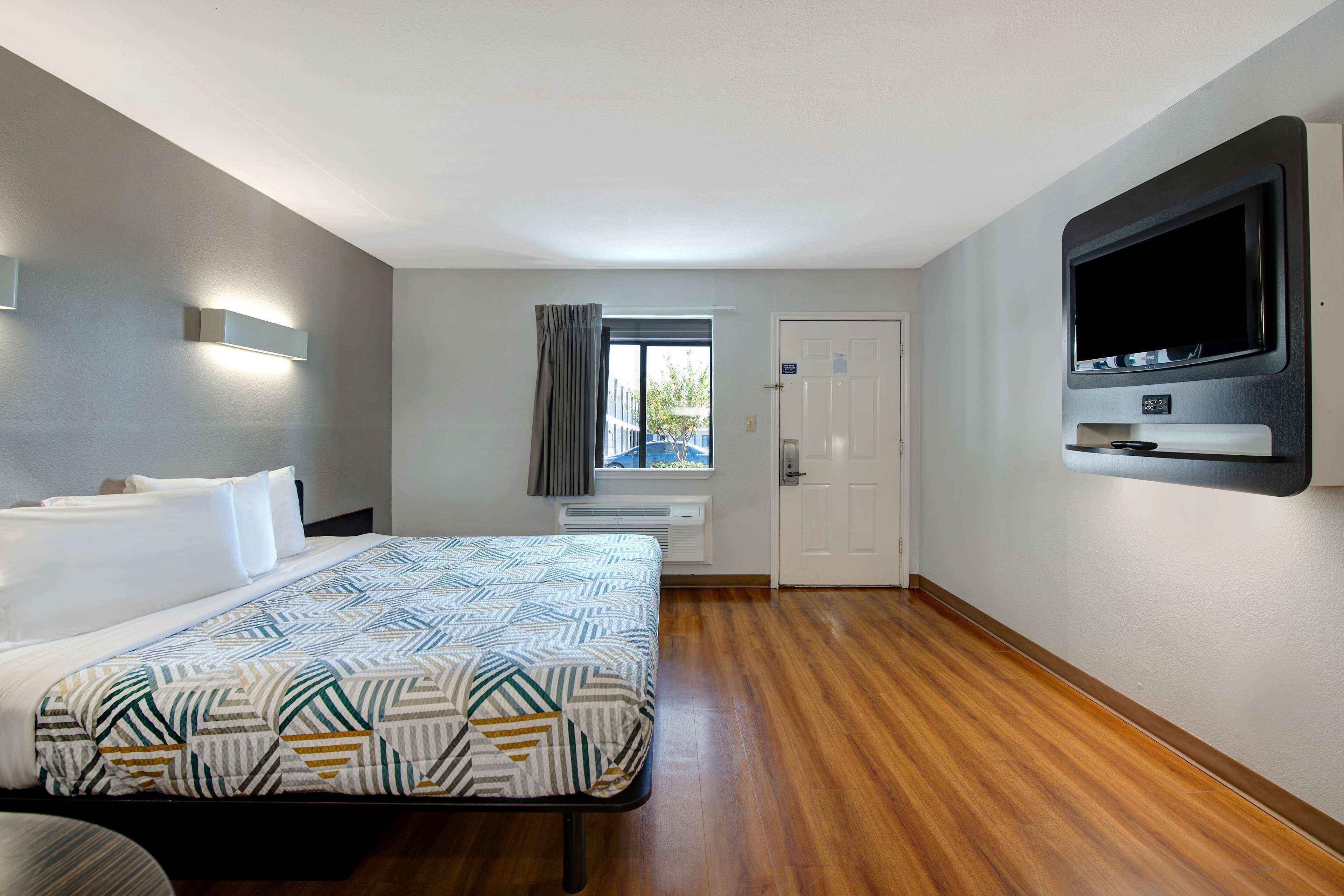 A hotel room with a bed, a TV mounted on the wall, a window with curtains, and a door.