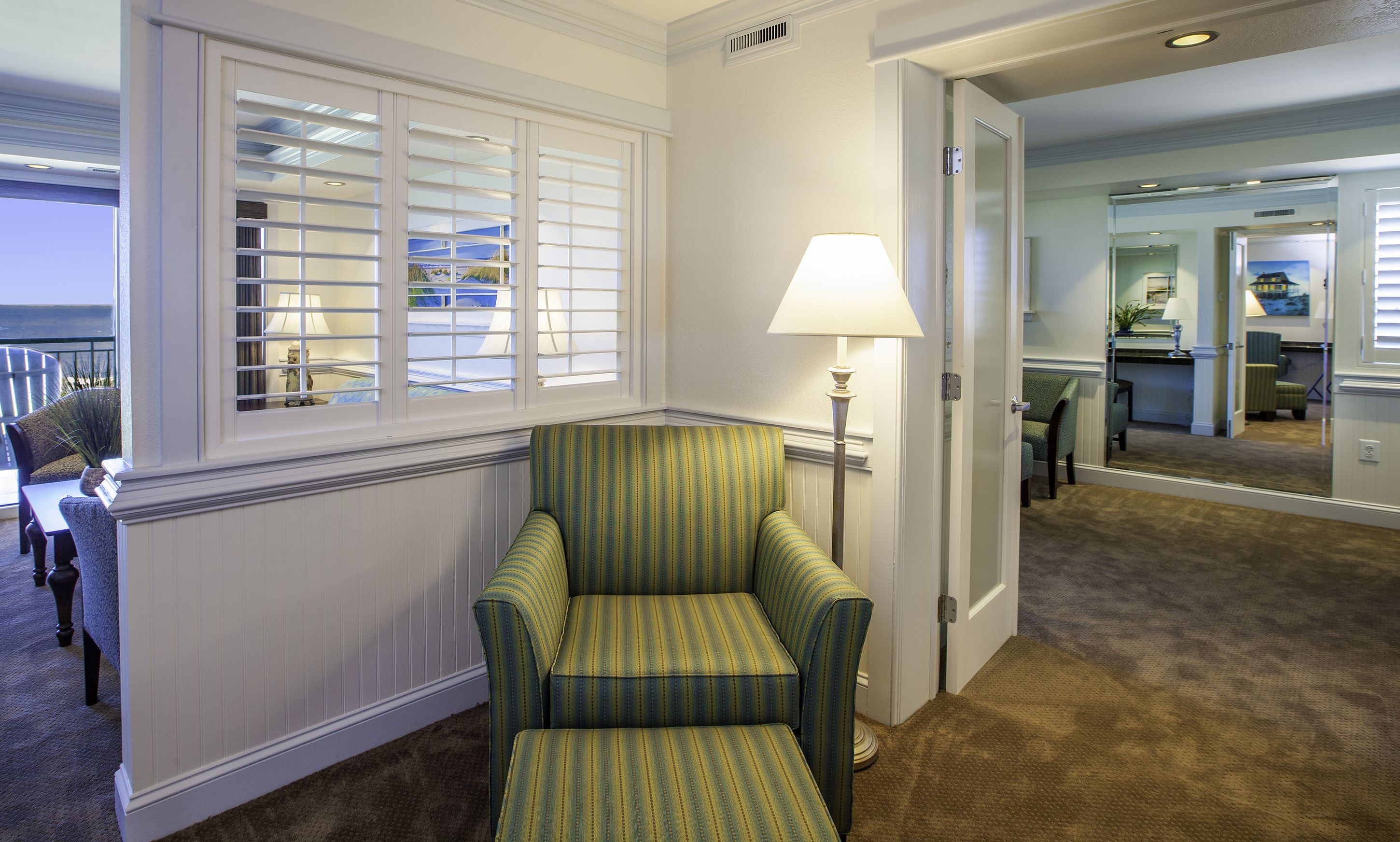 A hotel room with a green-striped armchair, a lamp, and a view through a window with blinds.