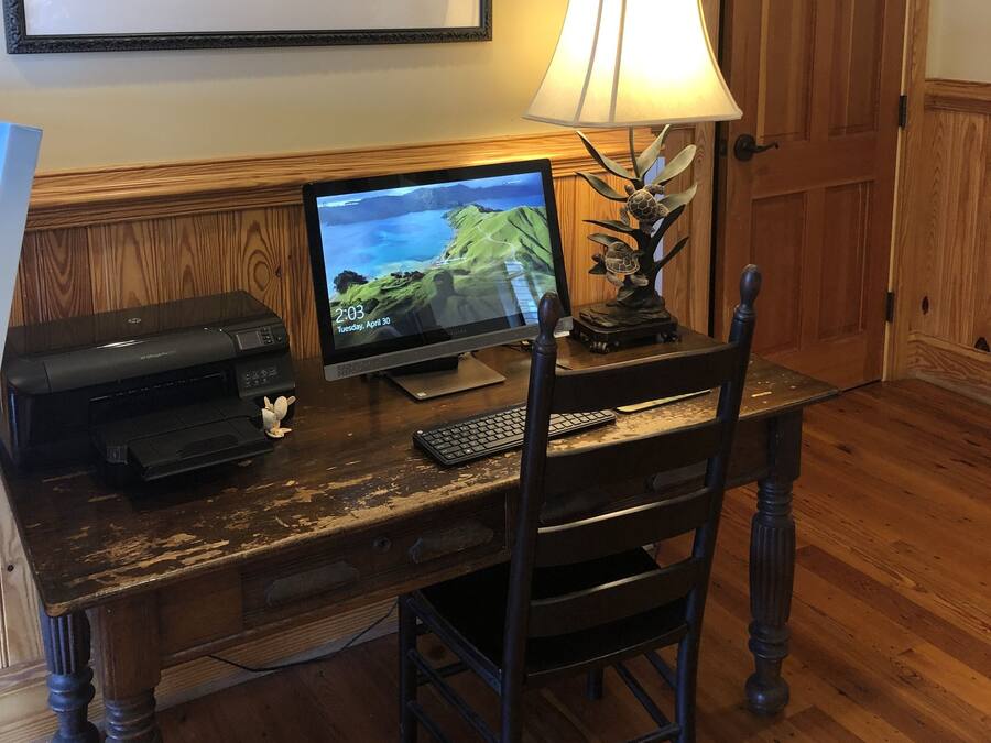 A wooden desk with a desktop computer, a lamp, and a printer. A framed portrait hangs on the wall above the desk.