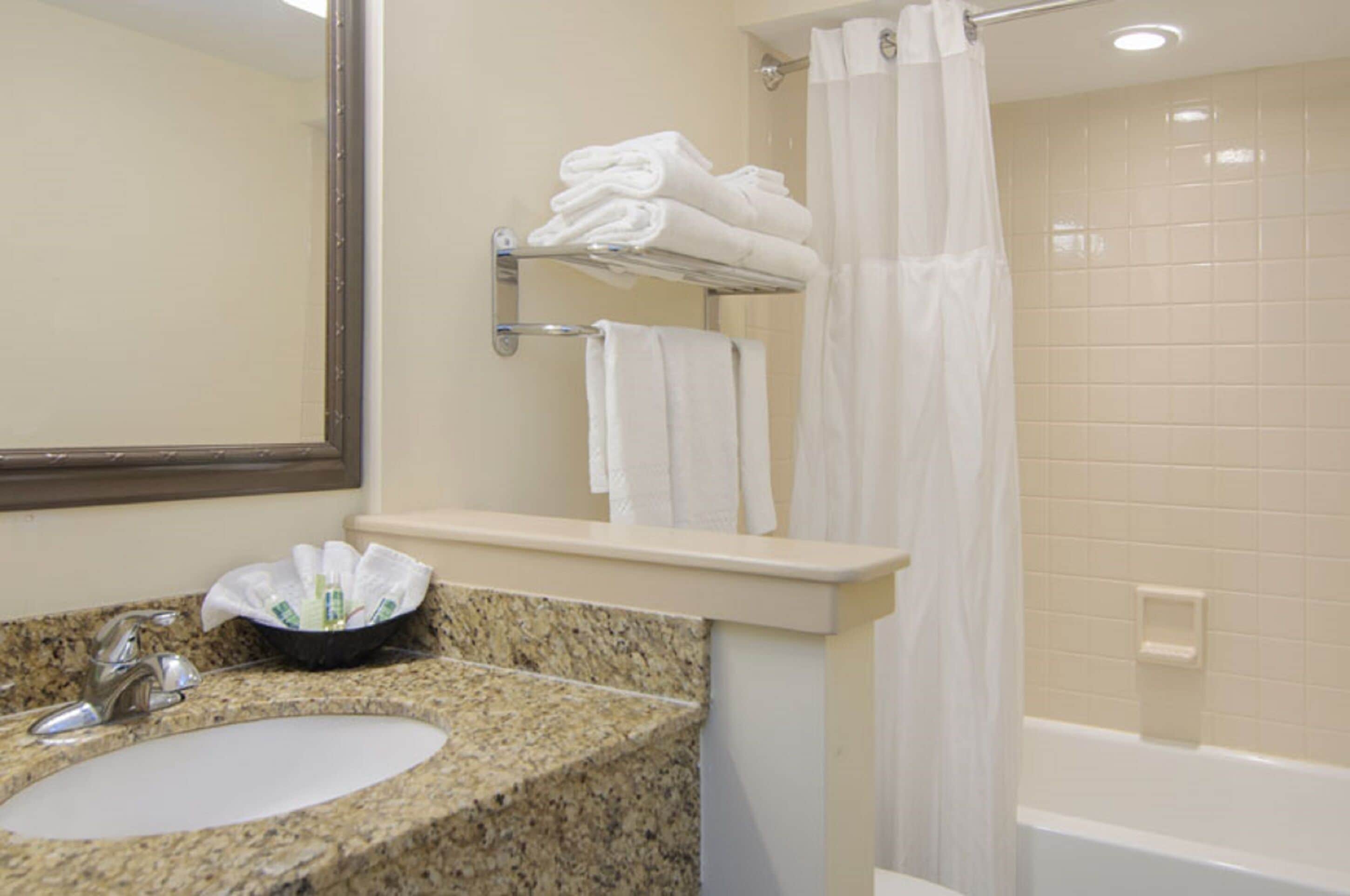 Une salle de bain avec un lavabo doté d’un plan de travail en granit, un miroir, un porte-serviettes avec des serviettes pliées et un rideau de douche.
