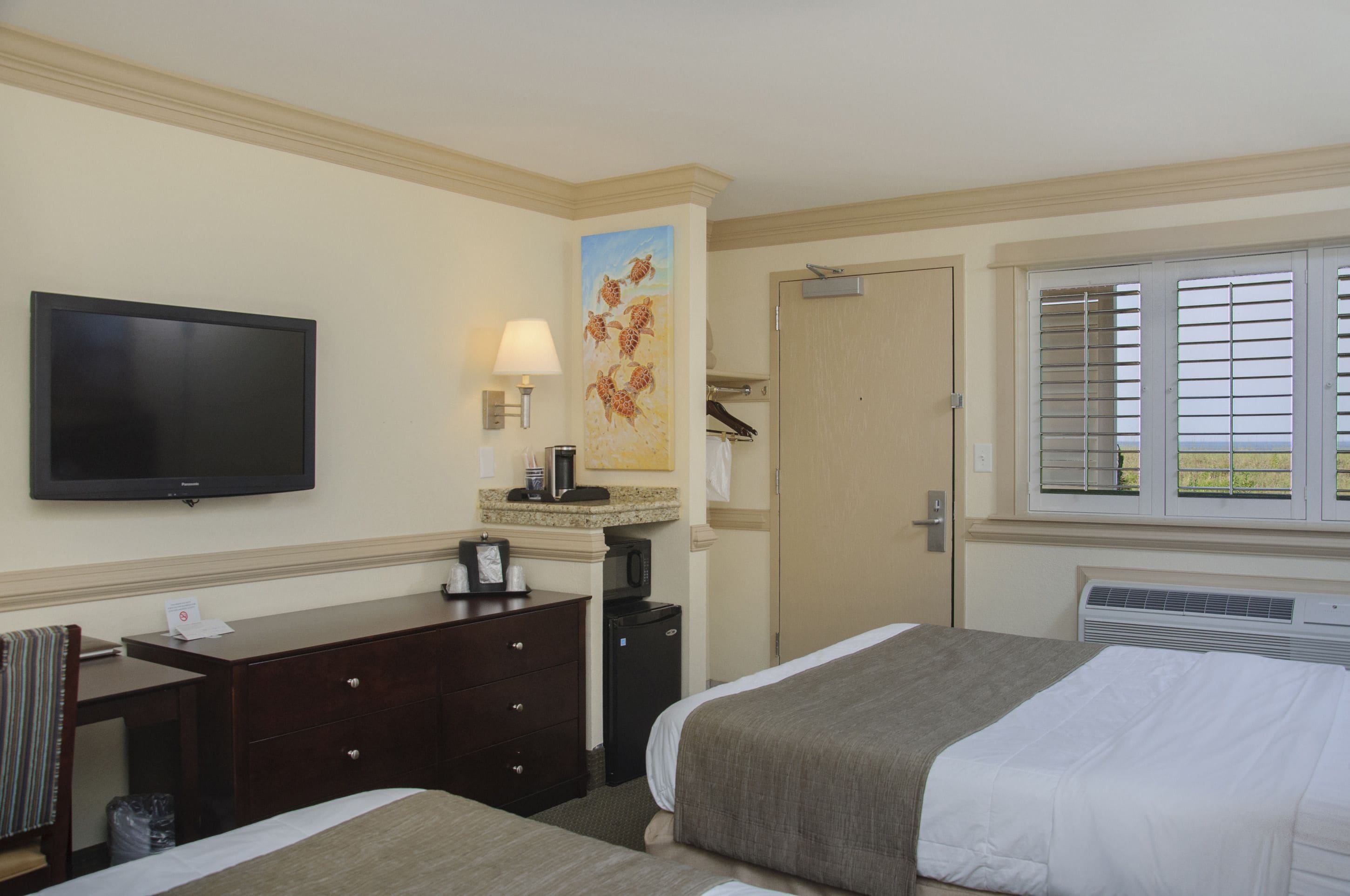 Habitación de hotel con cama, escritorio, televisor, un cuadro y ventana con persianas.