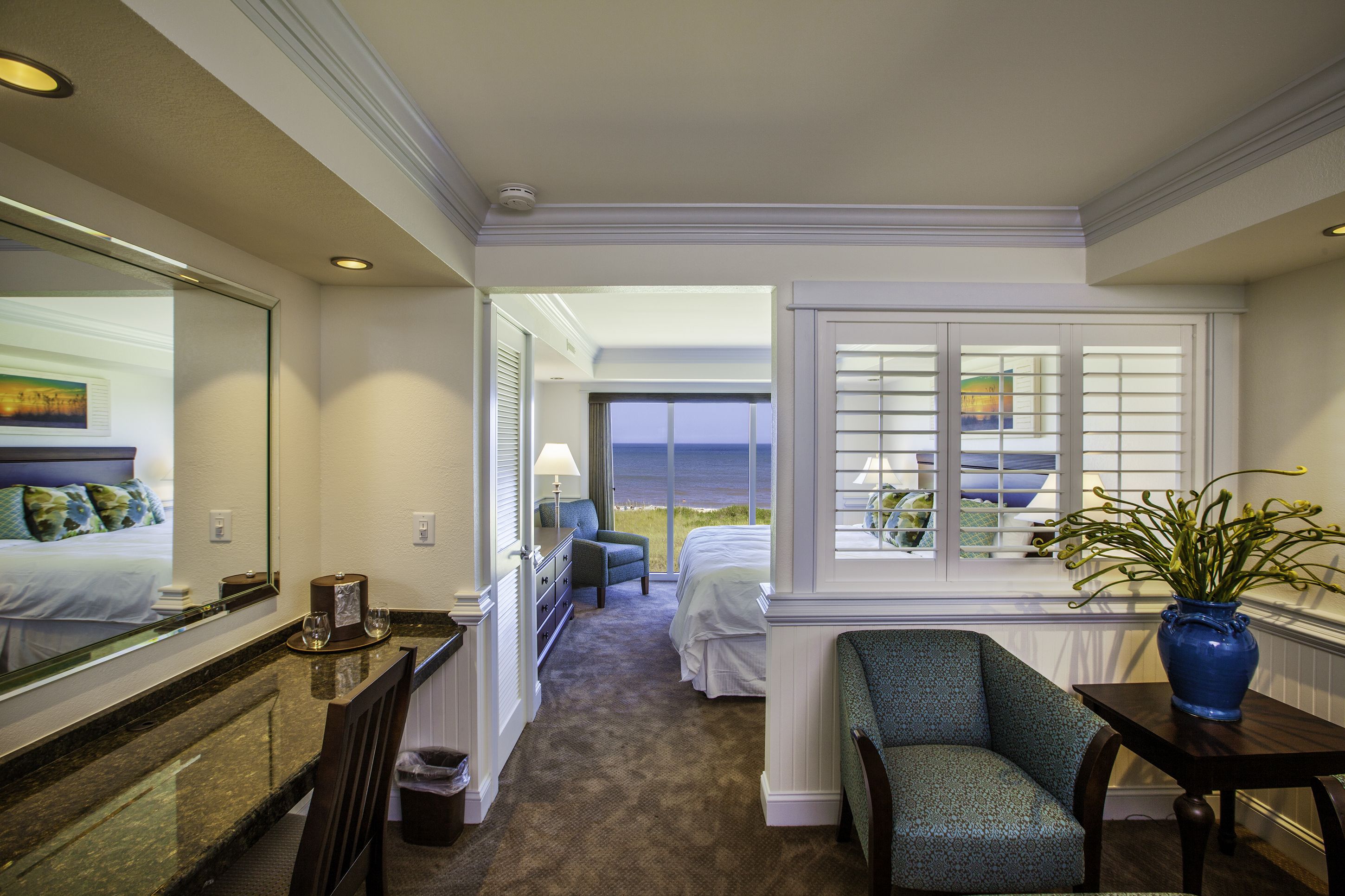 A hotel room with a bed, a chair, a table with a vase, a window with blinds, and a view of the ocean.
