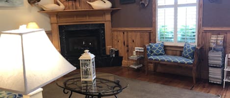 A cozy living room with a fireplace, a glass coffee table, a comfortable chair, and a lamp.