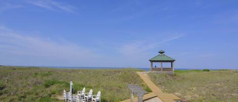 Ein Kiosk am Strand mit einer Holzterrasse, umgeben von grasbewachsenen Dünen und blauem Himmel.