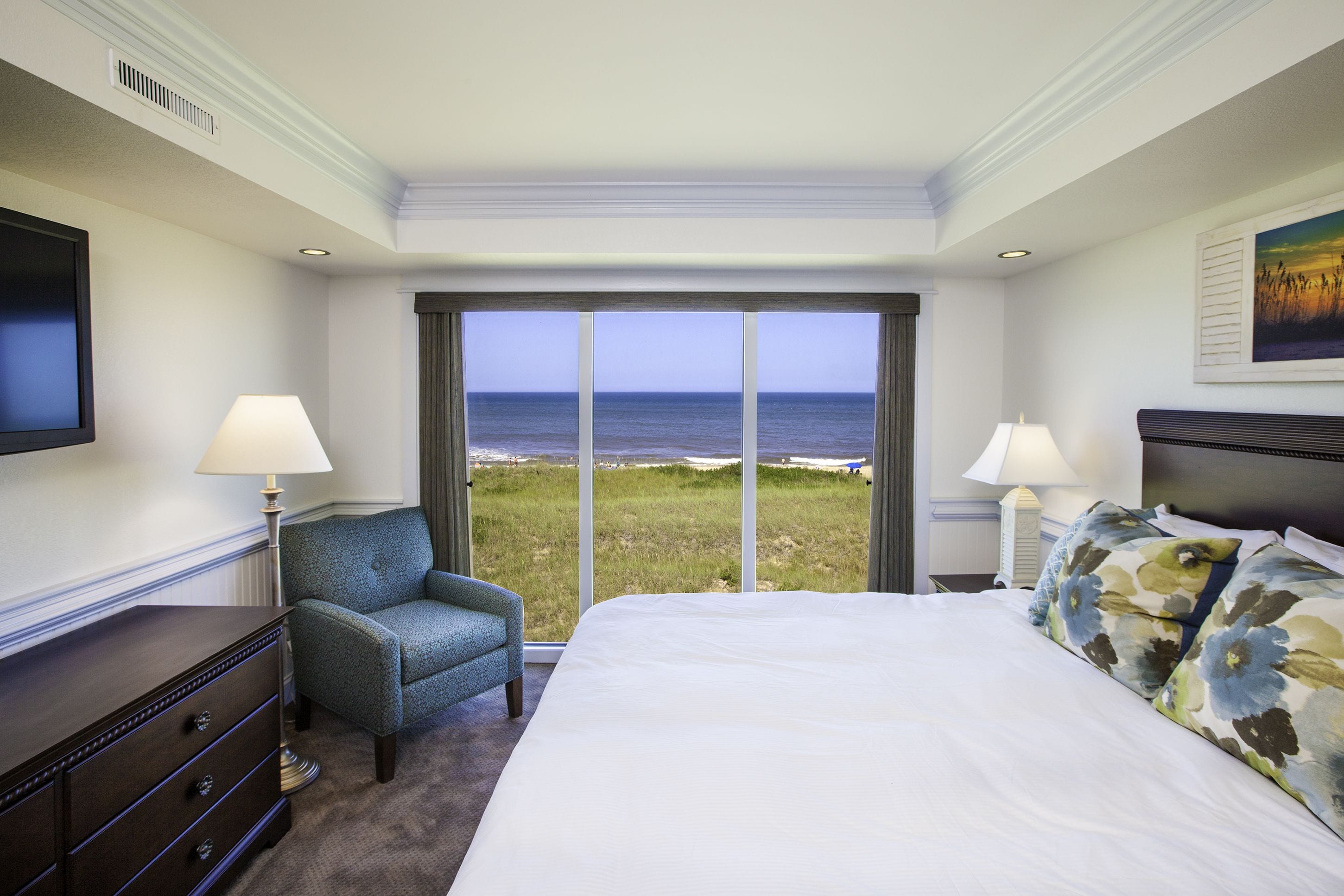 A hotel room with a large bed, two bedside lamps, a chair, a dresser, and a TV. The room has a large window with a view of the ocean.
