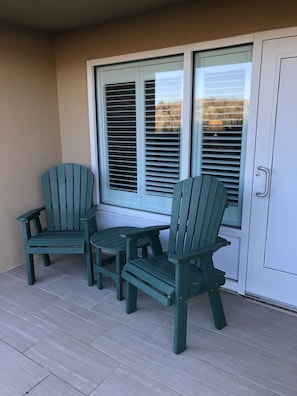 Un balcón con dos sillas Adirondack y una mesita, una puerta corrediza con persianas, y una puerta blanca con manija.