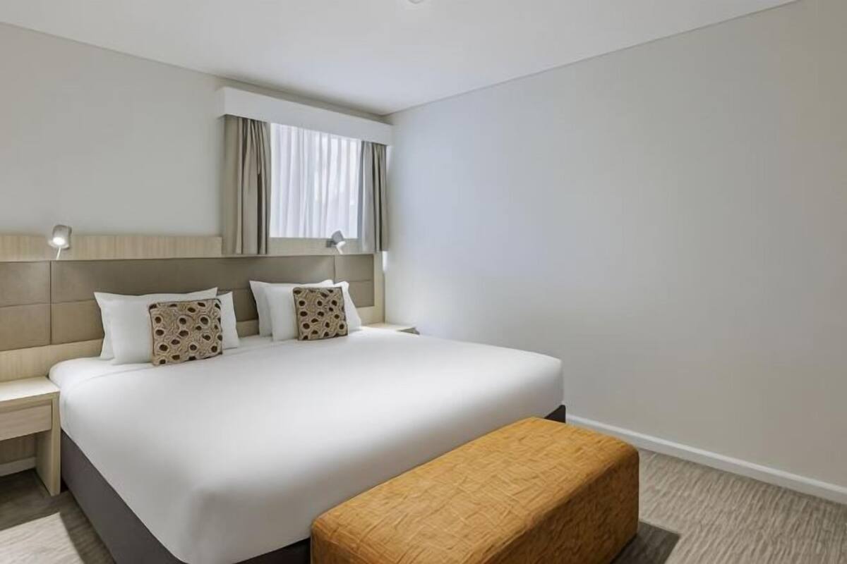 A hotel room with a large bed, bedside tables, a bench, a window with curtains, and a headboard.