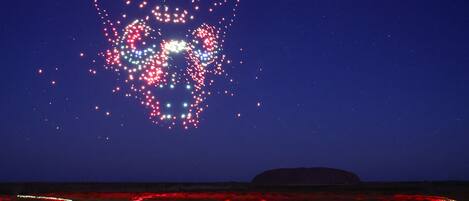 A night scene with a large illuminated structure resembling a heart shape in the sky, a flat landscape with red lighting, and a distant hill.