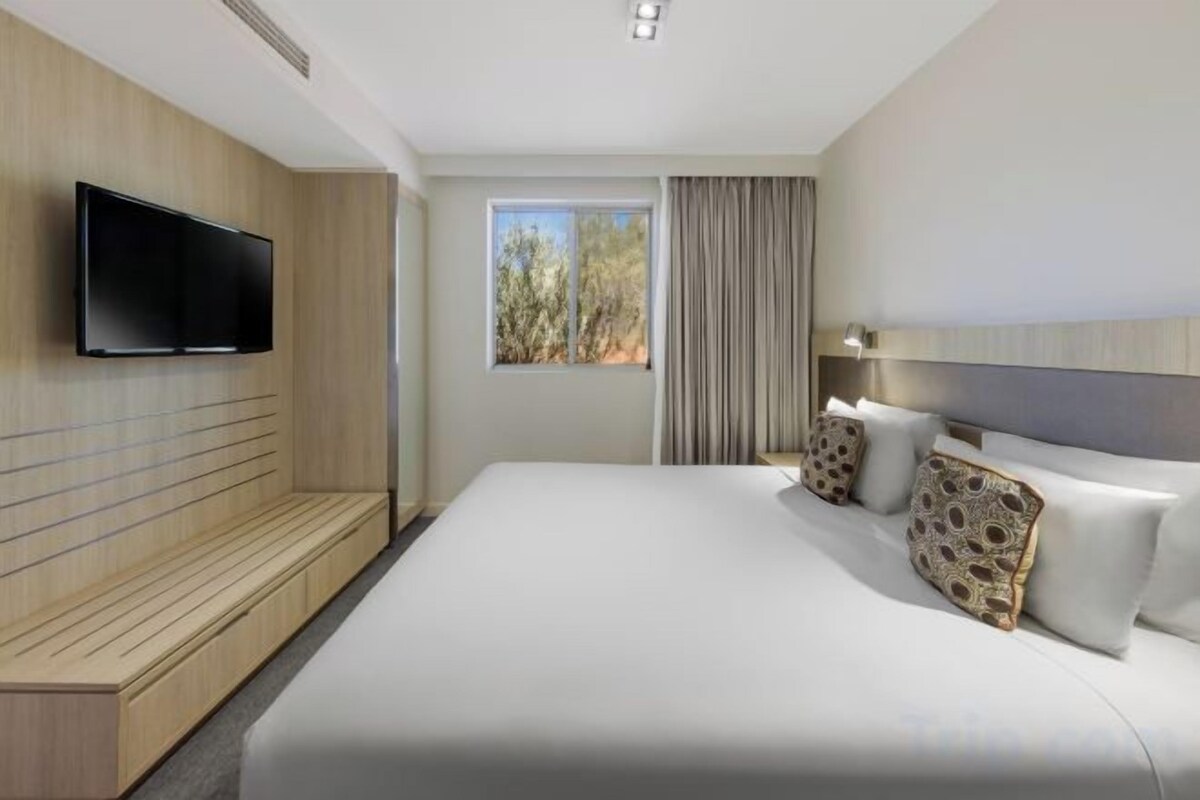 Hotellihuone, jossa on suuri sänky, seinälle asennettu taulutelevisio, ikkuna verhoilla sekä sängynpääty, jossa on valaisin.