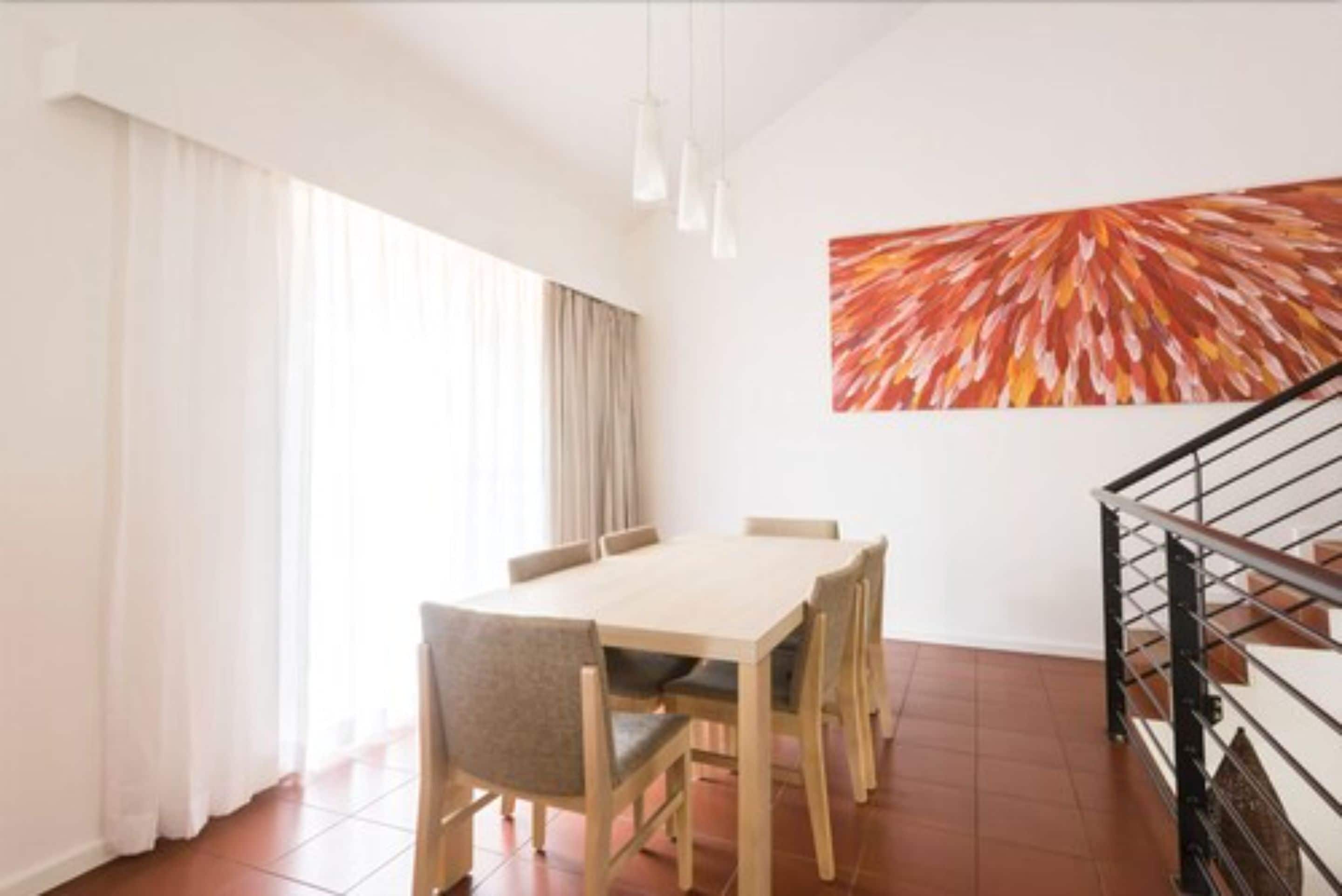A dining area with a wooden table and chairs, a large abstract painting on the wall, and a staircase with a metal railing.