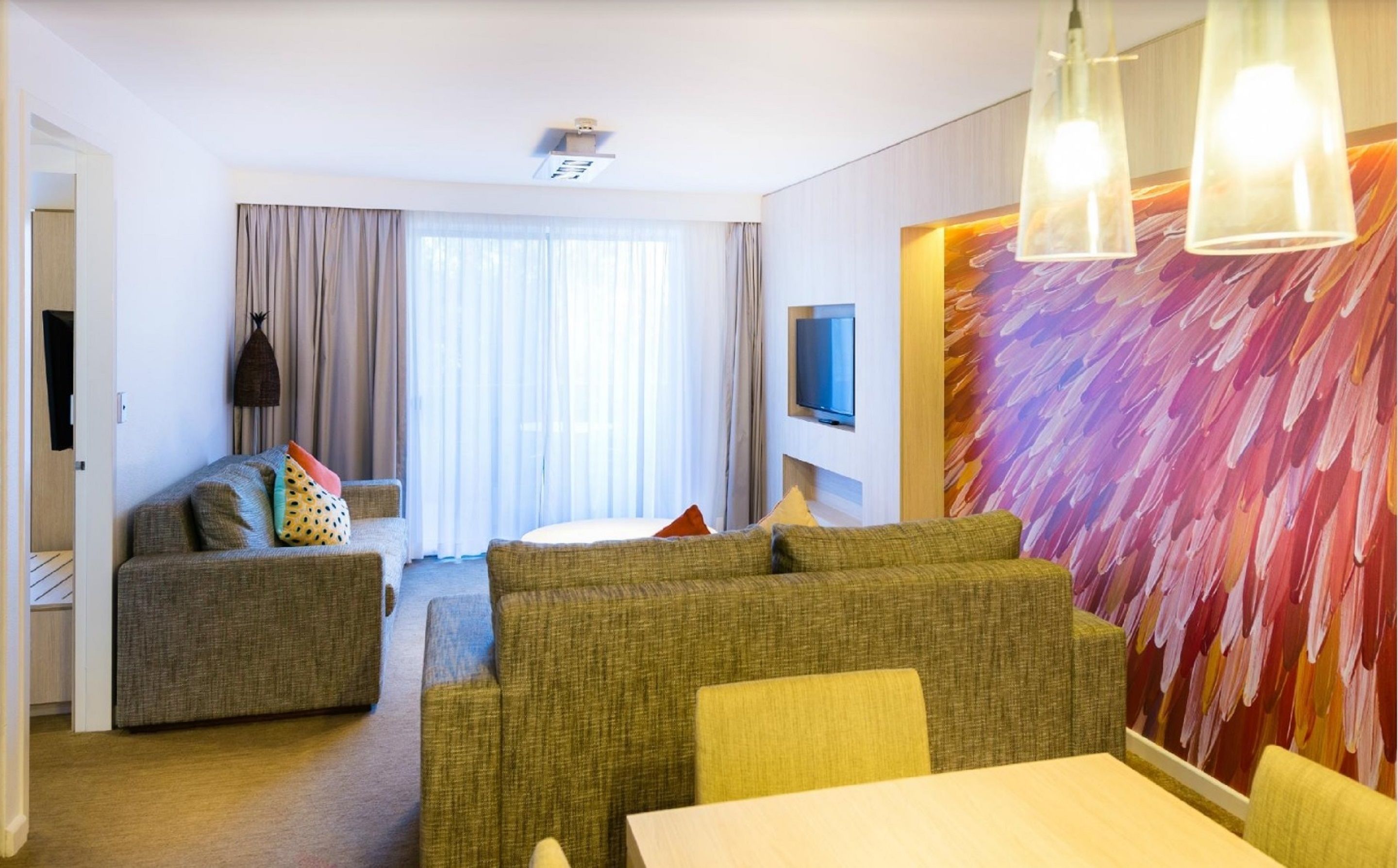 A modern hotel room with a sofa set, a dining table, a TV mounted on the wall, and a large abstract painting.