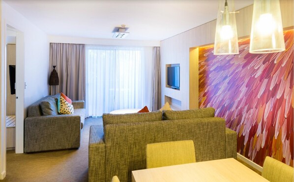 A modern hotel room with a sofa set, a dining table, a TV mounted on the wall, and a large abstract painting.