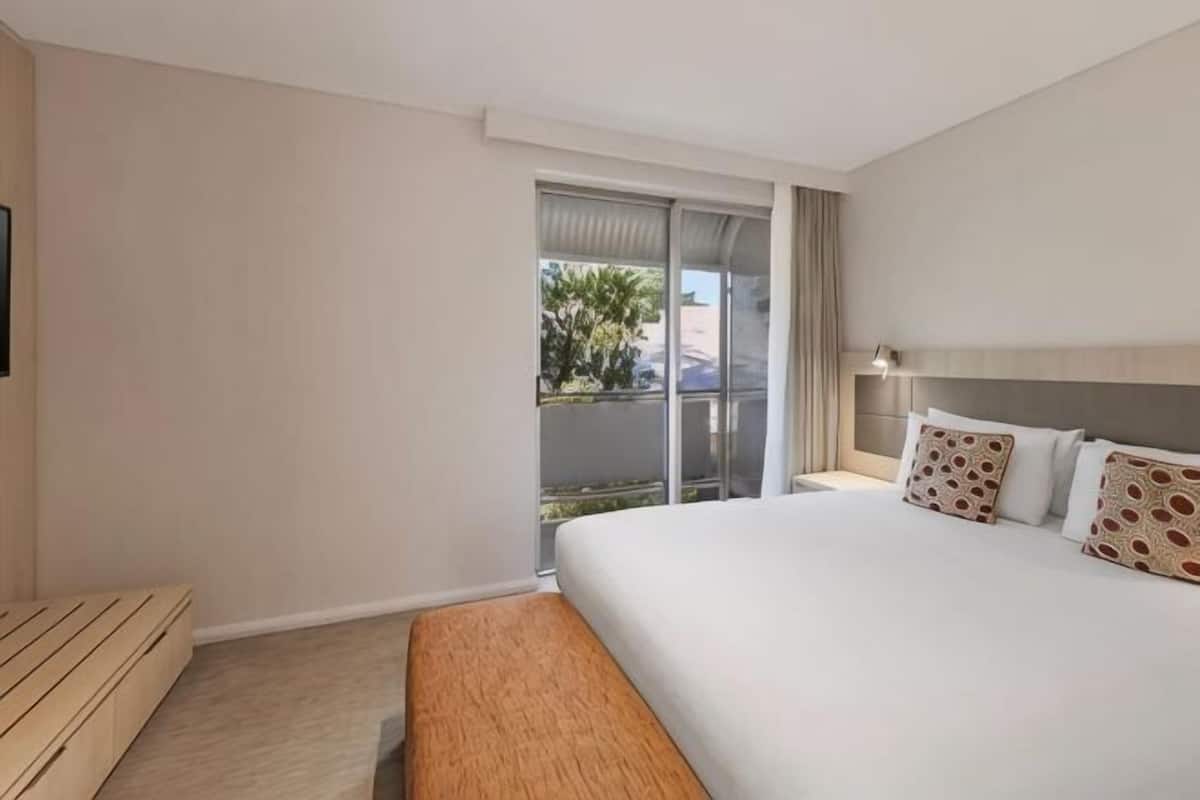 Habitación de hotel con cama, mesita de noche, lámpara y puerta corrediza de vidrio al exterior.