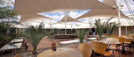 An outdoor patio area with round tables and chairs, shaded by large umbrellas, surrounded by palm trees.