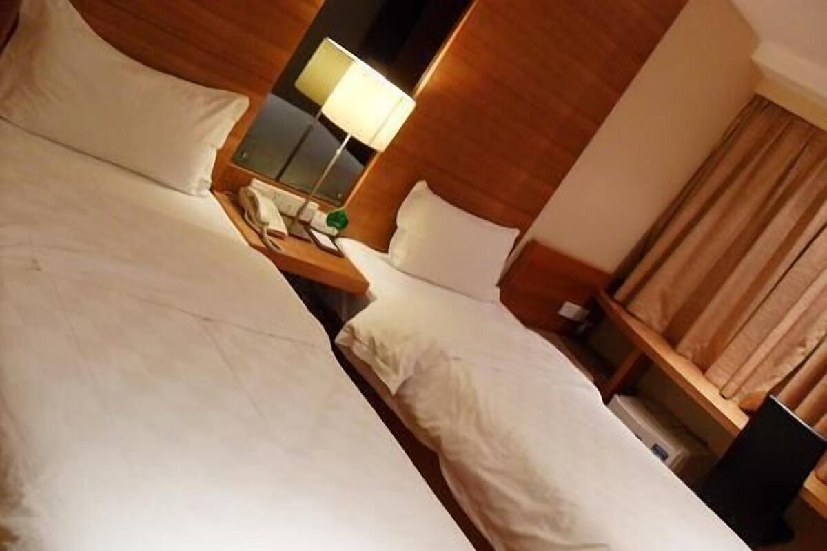 A hotel room with two beds, a bedside table with a lamp, and a window with curtains.