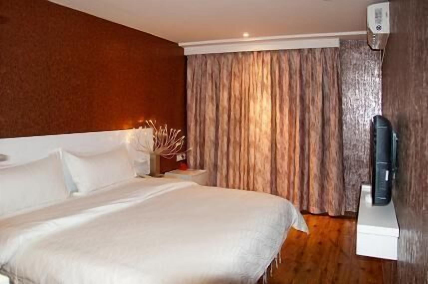 A hotel room with a large bed, a bedside table with a lamp, a TV mounted on the wall, and a window with curtains.