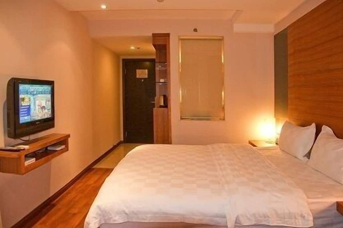 A hotel room with a bed, a TV on a stand, a wooden headboard, and a window with blinds.