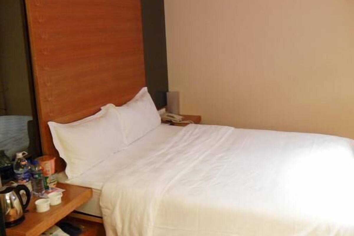 A hotel room with a bed, a wooden headboard, a nightstand with a phone, a coffee maker, and a cup.