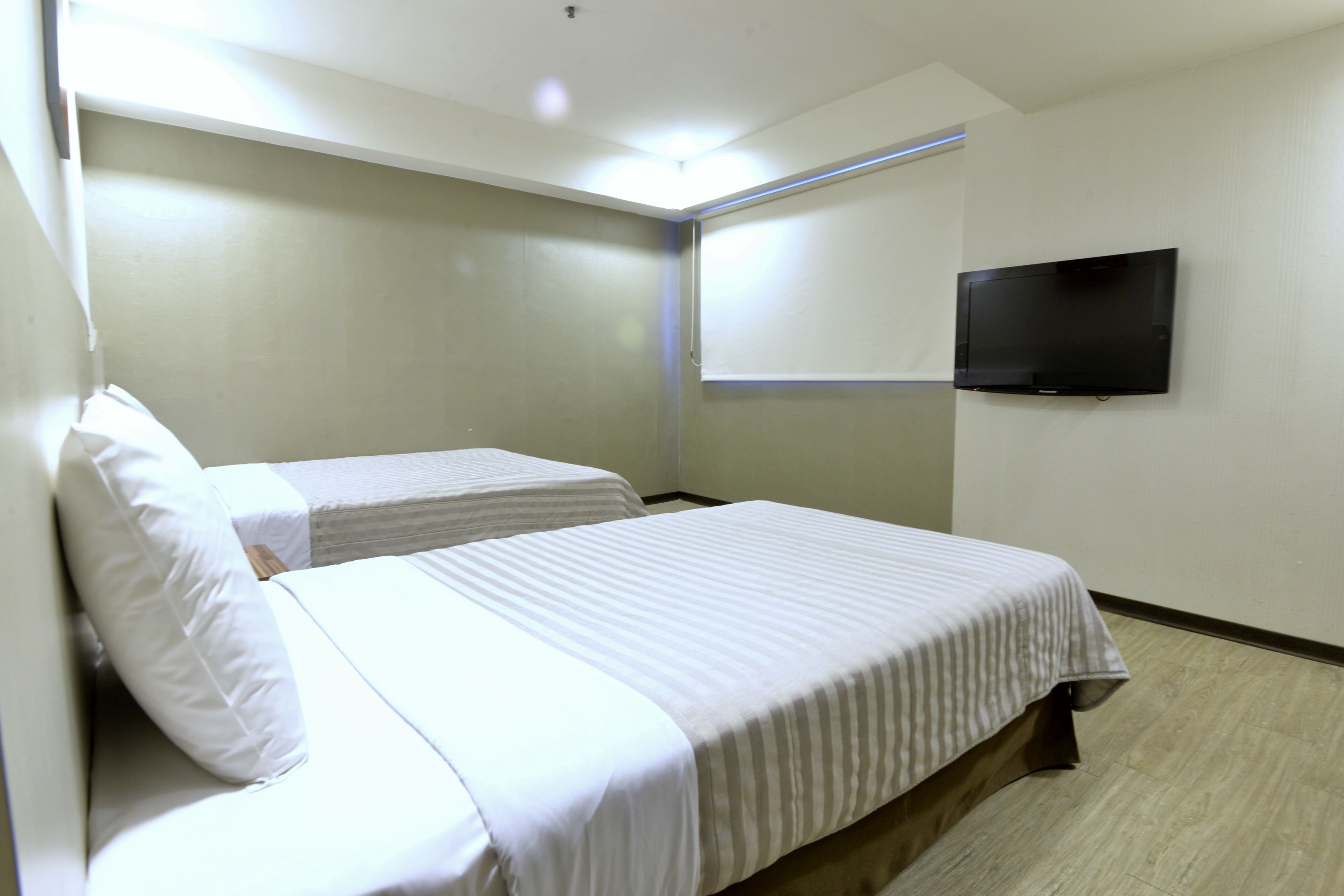 A hotel room with two single beds, a flat-screen TV mounted on the wall, and a window with blinds.