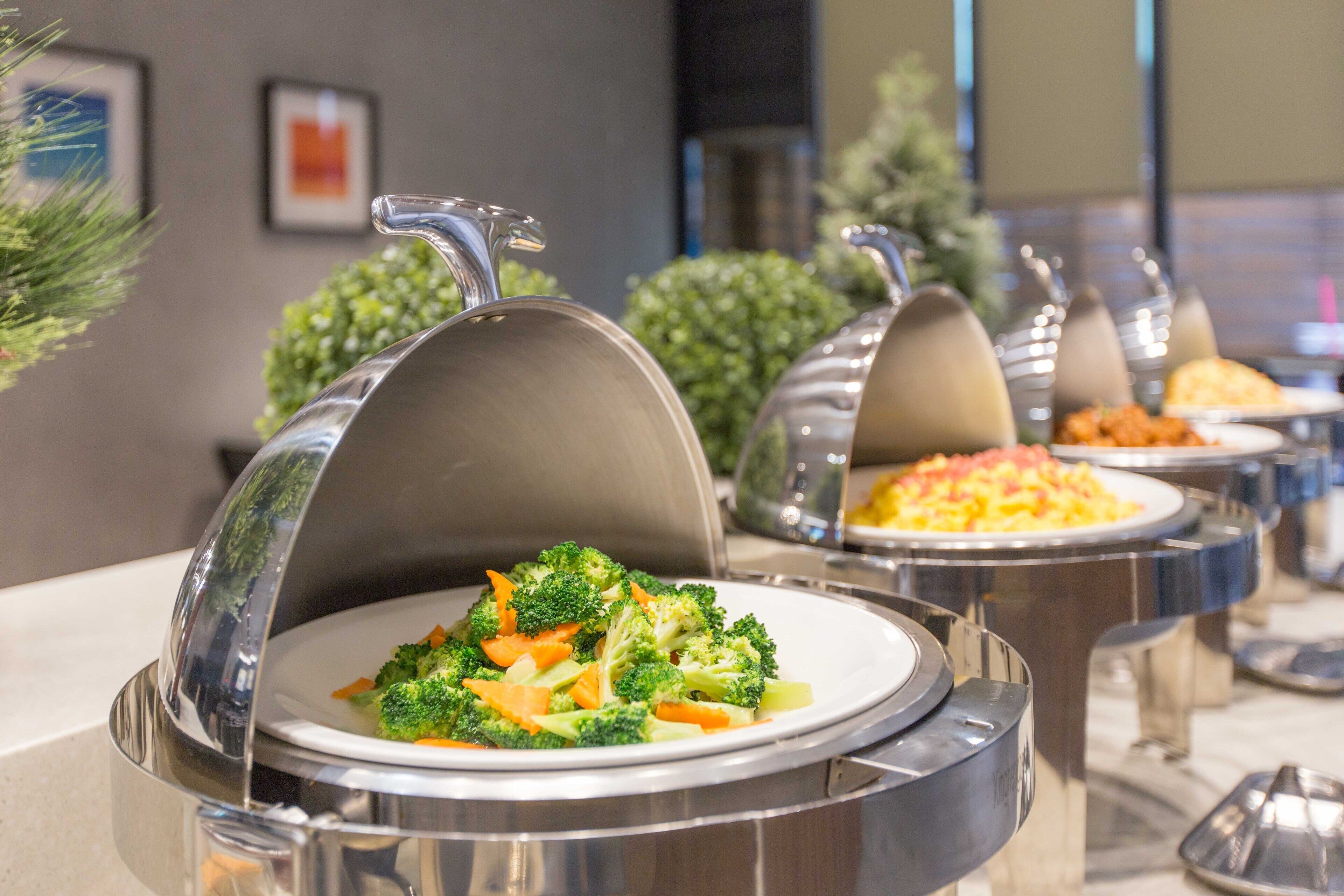 Servicio de comidas tipo buffet con recipientes cubiertos, incluyendo un plato de brócoli, en un entorno moderno con una paleta de colores neutros.