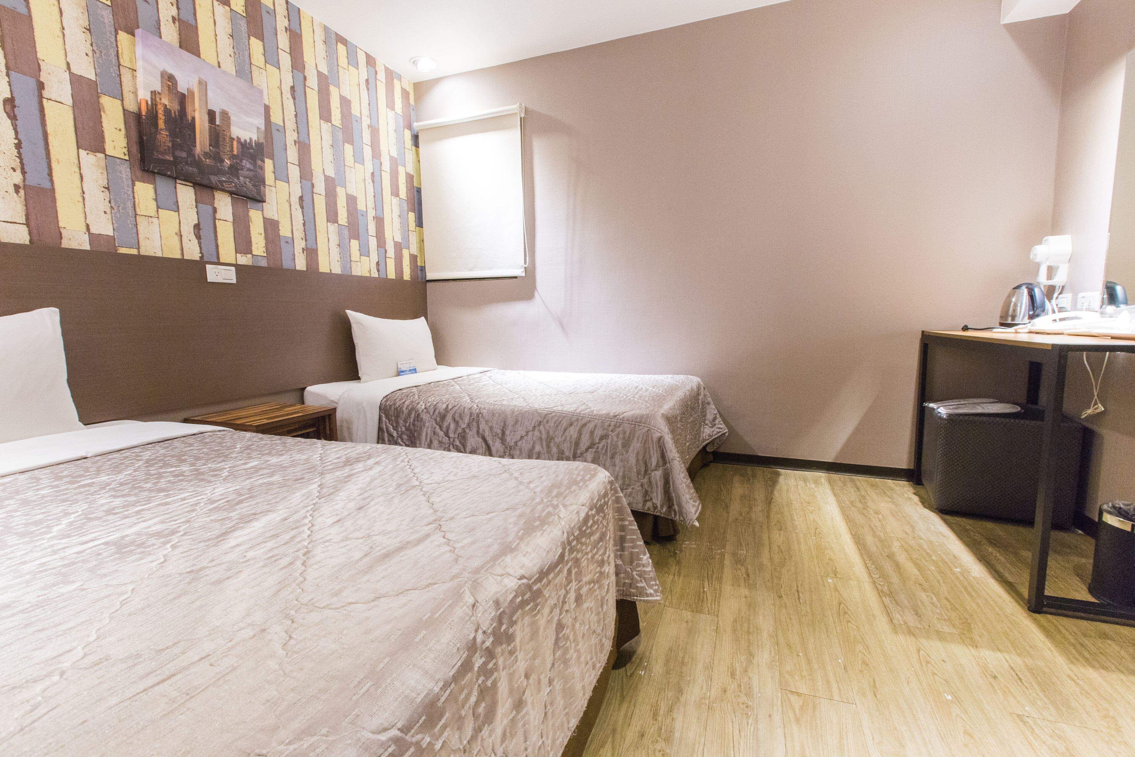 A hotel room with two beds, a small desk with a chair, a trash bin, and a window with a blind.