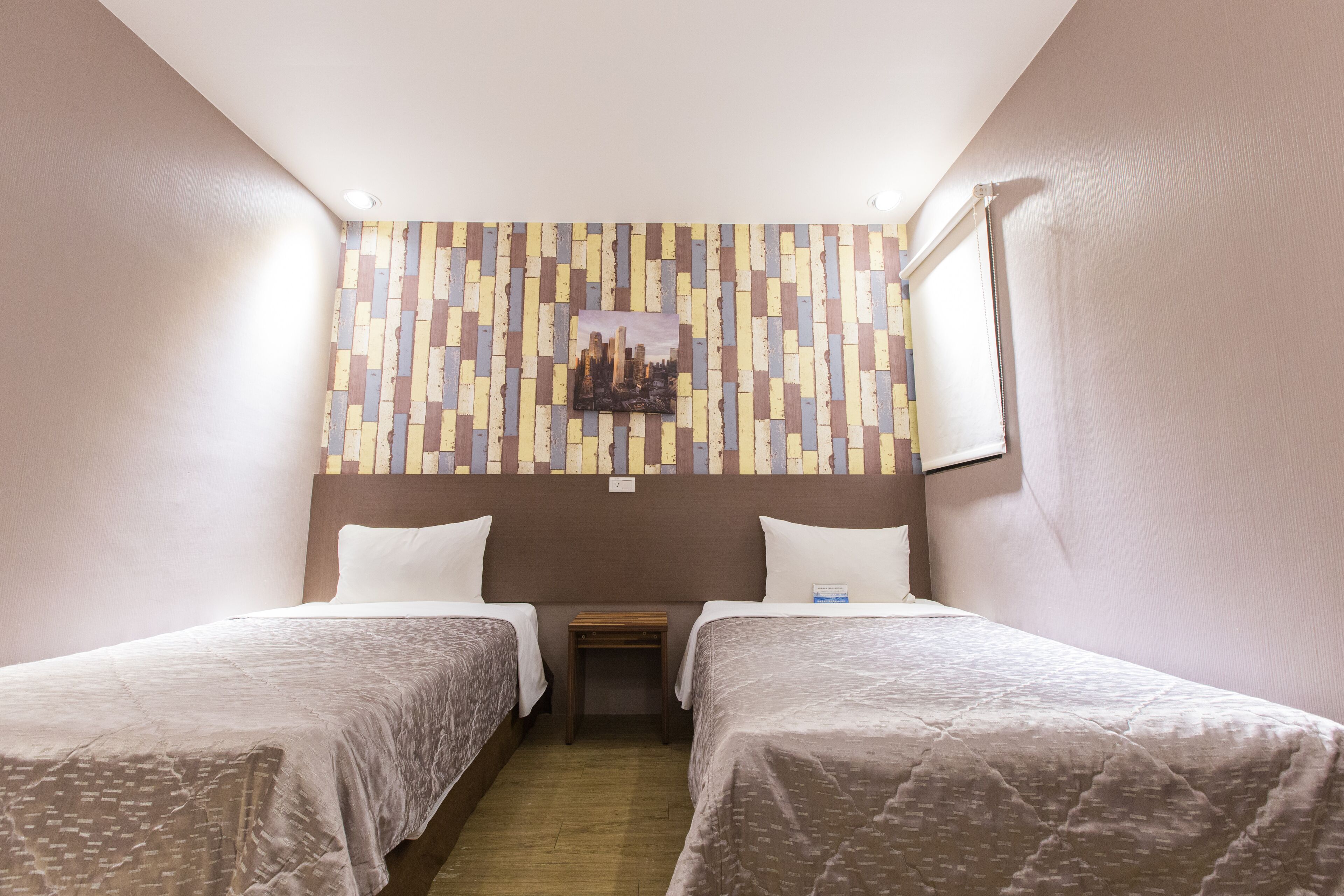 A hotel room with two single beds, a small wooden nightstand, a wall-mounted artwork, and a window with blinds.