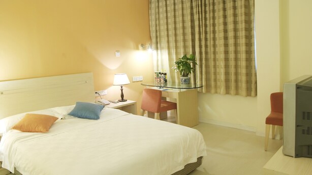 A hotel room with a bed, a desk with a lamp, a television, and a potted plant.