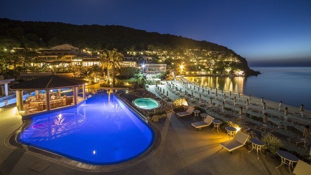 Vista nocturna de un complejo turístico con piscina, sillas de descanso y zona de playa.