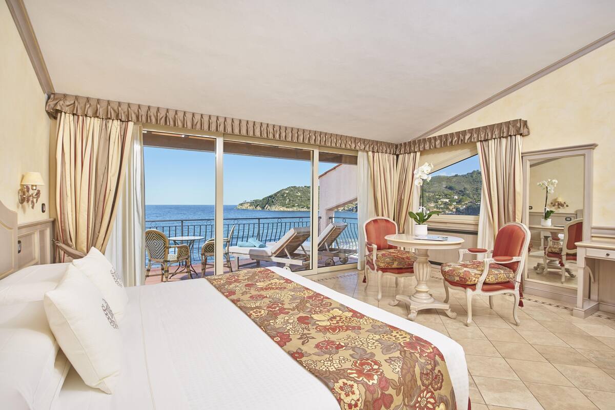 A hotel room with a bed, a desk, and a chair. There is a balcony with a view of the sea and a mountain.