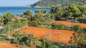 Una cancha de tenis con una vista panorámica al mar, rodeada de exuberante vegetación y una colina de fondo.