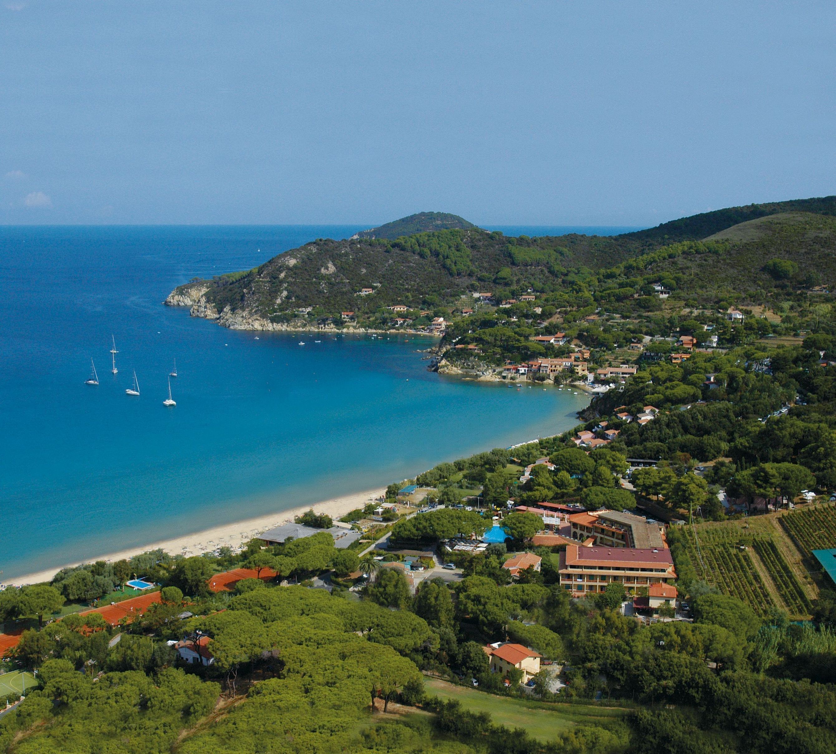 An aerial view of a coastal town with a beach, sailboats, and lush greenery.