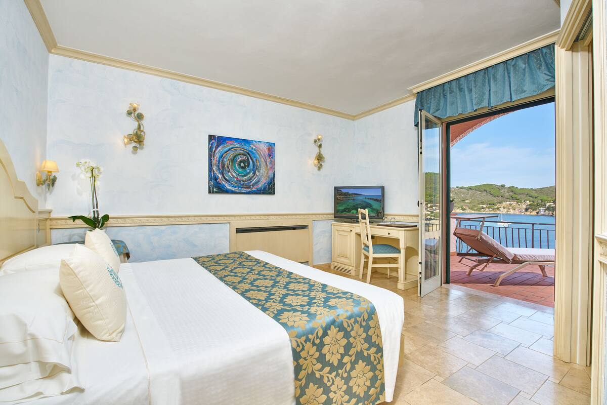 A hotel room with a bed, a desk, a chair, a TV, and a balcony with a view of the sea.