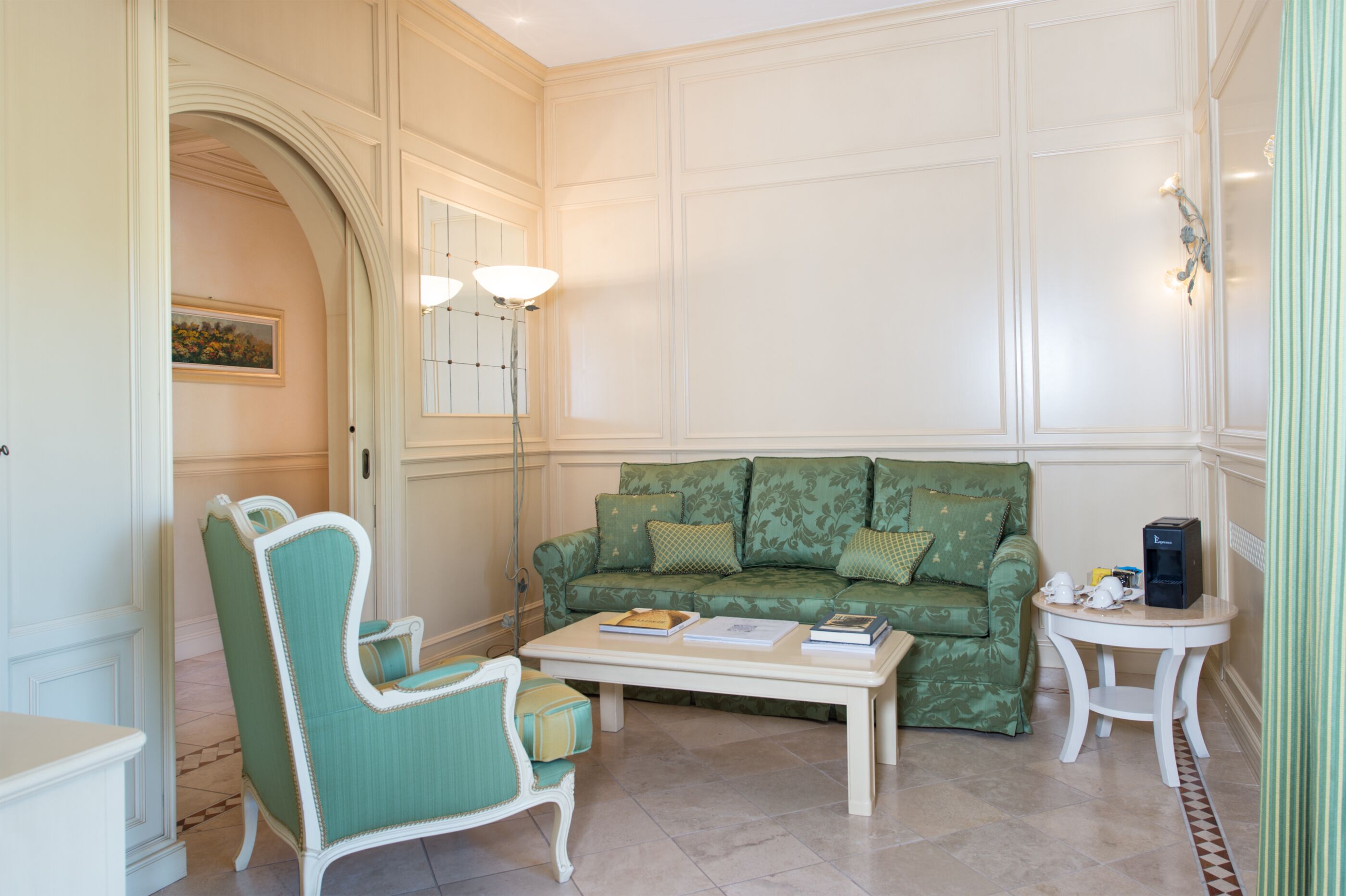 Una habitación con un sofá verde estampado, un sillón blanco, una mesita de centro con libros y una pequeña mesa redonda con una tetera.