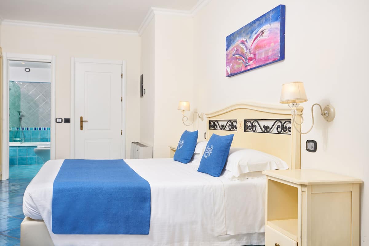 A hotel room with a bed, a nightstand, a lamp, a painting, and a bathroom visible through an open door.