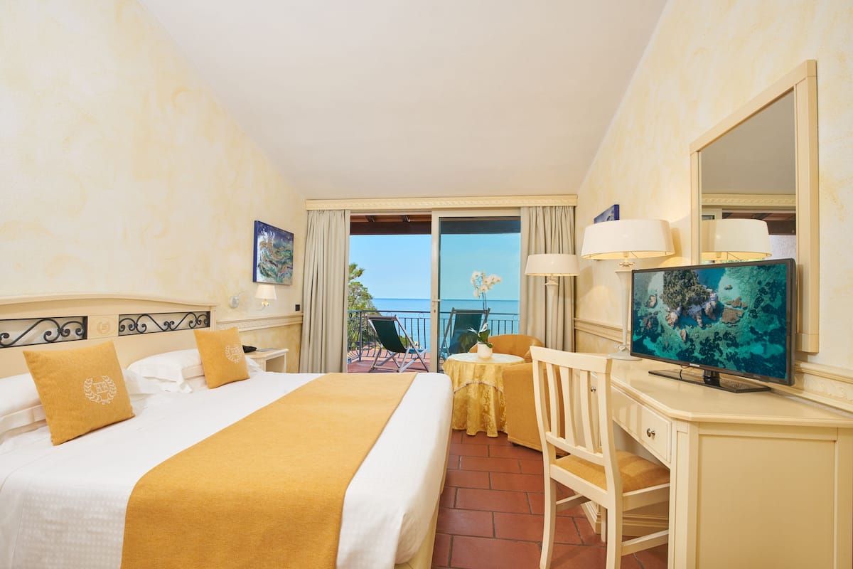 A hotel room with a bed, a desk, a TV, and a balcony with a view of the sea.