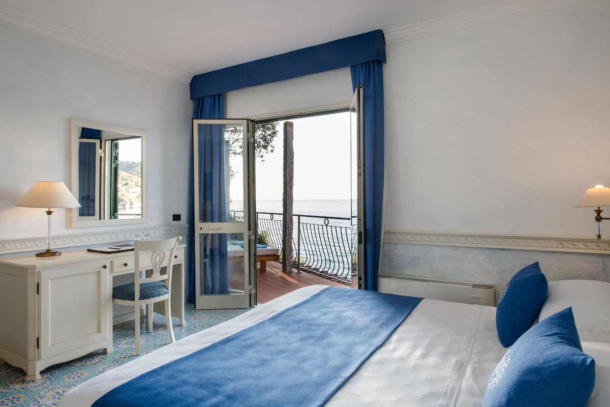 A hotel room with a bed, a desk, a chair, a balcony with a view of the sea, and a window with curtains.