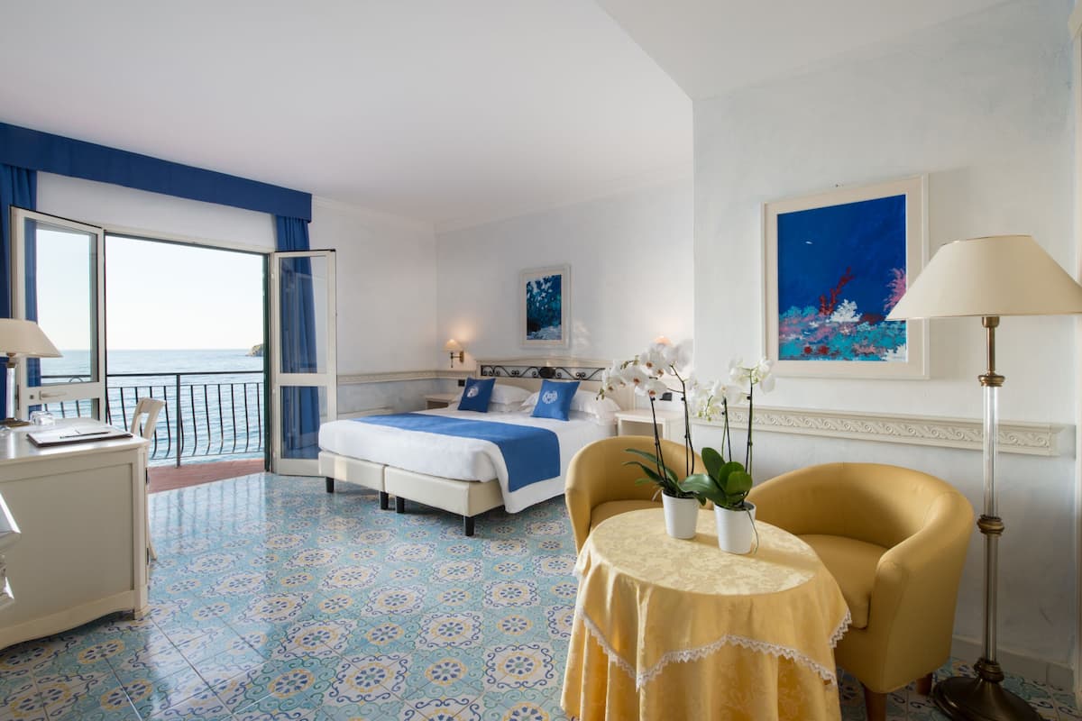 A hotel room with a bed, a desk, a chair, a table with a plant, and a painting on the wall.